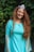A person with long, curly red hair is wearing a turquoise dress with sheer sleeves, standing in front of a lush, green leafy background. They are smiling and wearing a decorative headpiece with a blue gem.