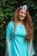 A person with long, curly red hair is wearing a turquoise dress with sheer sleeves, standing in front of a lush, green leafy background. They are smiling and wearing a decorative headpiece with a blue gem.