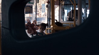 A scene inside a bus captured through a window. A child wearing a face mask is holding a colorful pinwheel. The bus's interior shows seats and metal support poles, and the outside appears bright with daylight visible through the windows.
