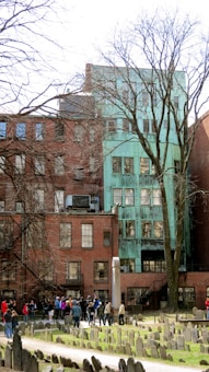 A group of people gather in a historic cemetery in front of old brick and green patina-covered buildings. Leafless trees are visible, and numerous gravestones are scattered across the grass.