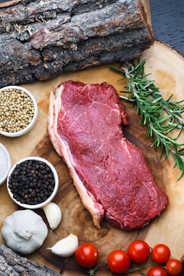 A raw steak is placed on a wooden surface surrounded by various food ingredients. On the right, a sprig of fresh rosemary rests beside the meat. To the left, small bowls contain coriander seeds and black pepper. Two cloves of garlic and several ripe cherry tomatoes add color to the scene. Pieces of rustic bark frame the top of the image.