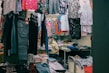 A small clothing shop filled with various garments hanging from racks. The shop includes colorful dresses, shirts, and pants, with neatly stacked clothes on shelves. A person wearing a mask is seated at the back beside these piled clothes, suggesting they are the shopkeeper.