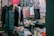 A small clothing shop filled with various garments hanging from racks. The shop includes colorful dresses, shirts, and pants, with neatly stacked clothes on shelves. A person wearing a mask is seated at the back beside these piled clothes, suggesting they are the shopkeeper.
