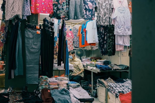 A small clothing shop filled with various garments hanging from racks. The shop includes colorful dresses, shirts, and pants, with neatly stacked clothes on shelves. A person wearing a mask is seated at the back beside these piled clothes, suggesting they are the shopkeeper.