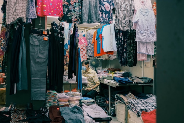 A small clothing shop filled with various garments hanging from racks. The shop includes colorful dresses, shirts, and pants, with neatly stacked clothes on shelves. A person wearing a mask is seated at the back beside these piled clothes, suggesting they are the shopkeeper.