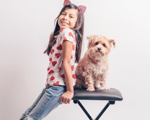 A young girl with long dark hair, wearing a heart-patterned shirt and sparkling cat ear headband, is leaning on a padded stool. Beside her sits a fluffy, tan-colored dog. Both the girl and the dog appear happy and relaxed, looking directly at the camera against a plain white background.