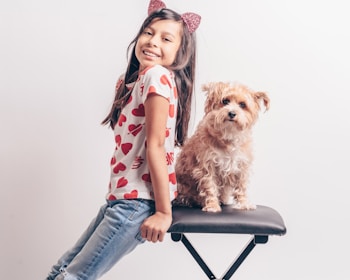 A young girl with long dark hair, wearing a heart-patterned shirt and sparkling cat ear headband, is leaning on a padded stool. Beside her sits a fluffy, tan-colored dog. Both the girl and the dog appear happy and relaxed, looking directly at the camera against a plain white background.
