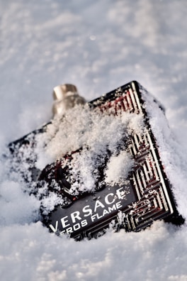 A perfume bottle partially covered in snow, with distinct branding and an intricate design. It is lying on a snowy surface, with some snowflakes clinging to its surface.