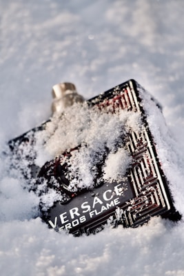 A perfume bottle partially covered in snow, with distinct branding and an intricate design. It is lying on a snowy surface, with some snowflakes clinging to its surface.