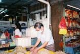 An indoor market scene with a man wearing a mask sorting dried seafood on a yellow table while a woman stands nearby, surrounded by hanging bags of goods and various market items.
