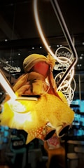 A doll wearing a yellow dress and hat sits on a lit surface, surrounded by various aesthetics including neon light shapes and a dimly lit, cozy background. The doll is adorned with a flower on its hat, giving it a playful and whimsical appearance.