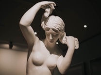 A classical marble statue of a woman with intricately detailed hair and calm, serene expression. The lighting highlights the smooth, polished surface and the subtle contours of the sculpture. The statue is posed with arms raised, hands adjusting her hair, set against a softly lit background.