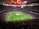 A large, packed stadium with bright lights illuminates a green soccer field. The scene is vibrant with a massive audience surrounding the pitch, visibly excited and engaged. The center of the field displays a prominent circular logo, suggesting a major sporting event. Players and staff can be seen on the field, likely preparing for a match.
