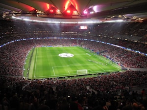 A large, packed stadium with bright lights illuminates a green soccer field. The scene is vibrant with a massive audience surrounding the pitch, visibly excited and engaged. The center of the field displays a prominent circular logo, suggesting a major sporting event. Players and staff can be seen on the field, likely preparing for a match.