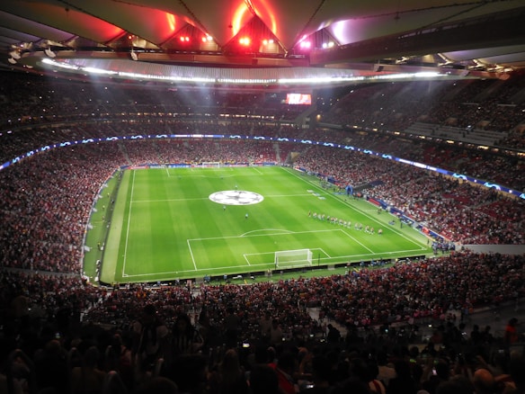 A large, packed stadium with bright lights illuminates a green soccer field. The scene is vibrant with a massive audience surrounding the pitch, visibly excited and engaged. The center of the field displays a prominent circular logo, suggesting a major sporting event. Players and staff can be seen on the field, likely preparing for a match.