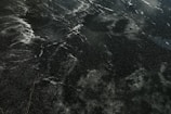A dark surface with an intricate pattern of white streaks and speckles, resembling ice or stone with natural veining and marbling.