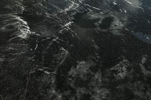 A dark surface with an intricate pattern of white streaks and speckles, resembling ice or stone with natural veining and marbling.