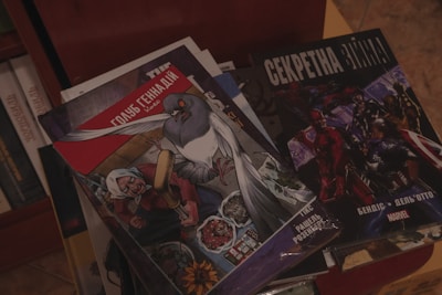 A collection of comic books is displayed on a surface. The cover of one comic features an illustration of a large pigeon and an old woman with various objects like food and flowers around them. Another comic book cover shows a group of superheroes in action poses.