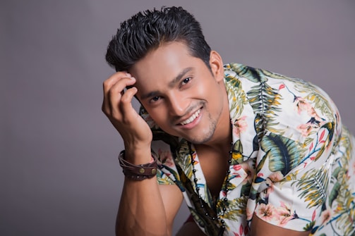 A smiling person with styled hair and a light beard is posing while wearing a tropical floral shirt. They have a bracelet on their wrist and are touching their hair in a playful manner, creating a friendly and approachable atmosphere.