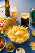 A variety of snacks and drinks are arranged on a blue surface. In the center, there is a glass bowl filled with potato chips with a bag of chips nearby. To the right, a can of beverage and a clear mug with dark beer are visible. On the left, an open bottle and a glass with light beer are placed, accompanied by a bottle cap. Two smaller bowls hold green olives and dip, likely yogurt-based, with a few pieces of crouton in one of the bowls. The setting suggests a casual snacking environment.