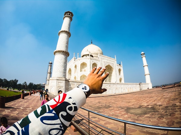 A close-up view of a hand wearing a colorful sleeve reaching towards the iconic white marble Taj Mahal, with its prominent domes and minarets against a clear blue sky. The scene includes a walkway bordered by grass and several visitors walking around the complex.