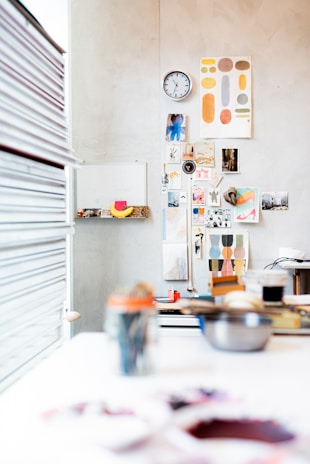 A creative workspace with a wall adorned with various art pieces, photographs, and a clock. The foreground shows blurred art supplies and tools scattered on a white table. Bright and colorful prints add vibrancy, and a stack of books is visible to the left.