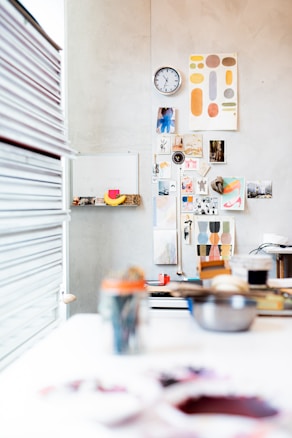 A creative workspace with a wall adorned with various art pieces, photographs, and a clock. The foreground shows blurred art supplies and tools scattered on a white table. Bright and colorful prints add vibrancy, and a stack of books is visible to the left.