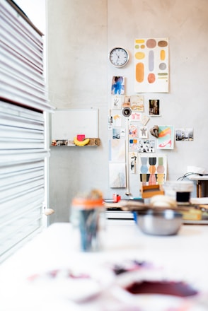 A creative workspace with a wall adorned with various art pieces, photographs, and a clock. The foreground shows blurred art supplies and tools scattered on a white table. Bright and colorful prints add vibrancy, and a stack of books is visible to the left.