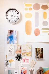 A wall features a silver-rimmed clock displaying a time around 10:10. Below the clock are various decorative art pieces and photographs pinned to the wall. These include abstract images, illustrations of animals, and photographs of nature scenes. A color palette poster with different shades and shapes is displayed prominently.