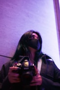 A person with long dark hair wears a black mask and a dark coat, holding a camera while looking upward. The background is softly illuminated with purple light, giving the scene a modern, artistic vibe.