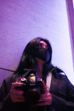 A person with long dark hair wears a black mask and a dark coat, holding a camera while looking upward. The background is softly illuminated with purple light, giving the scene a modern, artistic vibe.