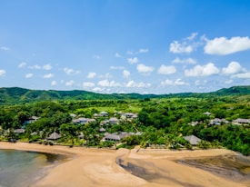 A picturesque landscape featuring a sandy beach bordered by clear waters. Lush green vegetation and hills form a verdant backdrop. Several small structures, possibly houses or villas, are scattered among the greenery. White, fluffy clouds float across a bright blue sky, enhancing the serene atmosphere.
