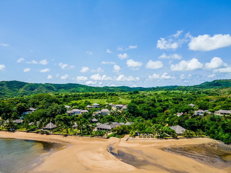 A picturesque landscape featuring a sandy beach bordered by clear waters. Lush green vegetation and hills form a verdant backdrop. Several small structures, possibly houses or villas, are scattered among the greenery. White, fluffy clouds float across a bright blue sky, enhancing the serene atmosphere.
