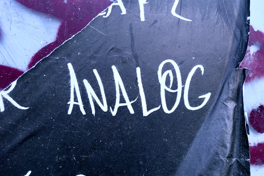The word 'ANALOG' is written in white, bold letters on a dark, textured surface with a slight tear on the side. The background has a mix of maroon and black colors with some visible scratches.