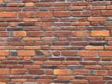 A pattern of red and orange bricks arranged in a horizontal layout, featuring variations in texture and shade. The mortar lines between the bricks are visible and uniform, providing a consistent and orderly appearance.