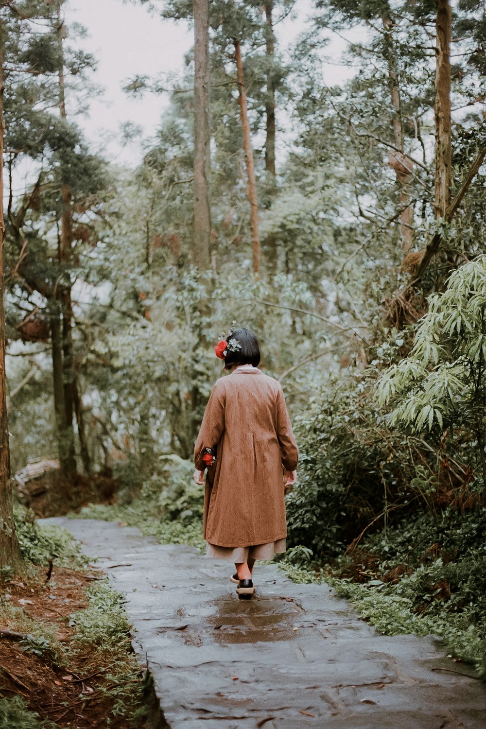 donna in cappotto marrone che cammina sul sentiero tra gli alberi durante il giorno