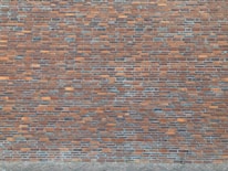 A brick wall composed of numerous rectangular bricks, displaying a mix of reddish-brown and gray tones. The bricks are neatly arranged in a horizontal pattern, creating a textured surface. The lower section features a cobblestone pavement.