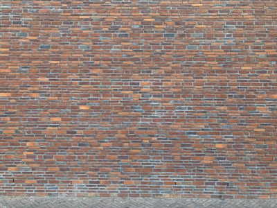 A brick wall composed of numerous rectangular bricks, displaying a mix of reddish-brown and gray tones. The bricks are neatly arranged in a horizontal pattern, creating a textured surface. The lower section features a cobblestone pavement.