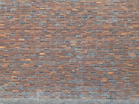 A brick wall composed of numerous rectangular bricks, displaying a mix of reddish-brown and gray tones. The bricks are neatly arranged in a horizontal pattern, creating a textured surface. The lower section features a cobblestone pavement.