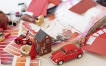 A creative workspace featuring various art supplies and items arranged on a table. There are color swatches, watercolor paints, and open sketchbooks displaying floral and abstract designs. A small red toy car, a model house, a spool of tape, and a miniature gnome add whimsical elements. Paint tubes and brushes are scattered, suggesting an artistic process in progress. The overall scene is vibrant and cluttered.