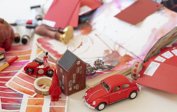A creative workspace featuring various art supplies and items arranged on a table. There are color swatches, watercolor paints, and open sketchbooks displaying floral and abstract designs. A small red toy car, a model house, a spool of tape, and a miniature gnome add whimsical elements. Paint tubes and brushes are scattered, suggesting an artistic process in progress. The overall scene is vibrant and cluttered.