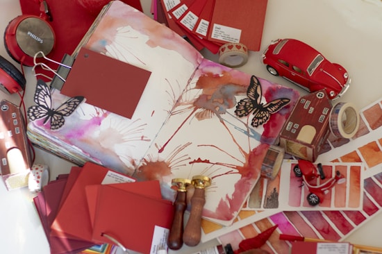 An open sketchbook features watercolor art with splashes of red, orange, and pink hues. Surrounding the sketchbook are various art and lifestyle objects, including red headphones, paint color swatches, a miniature red car, butterfly cutouts, paint swatches, a model house, and brass wax seals.