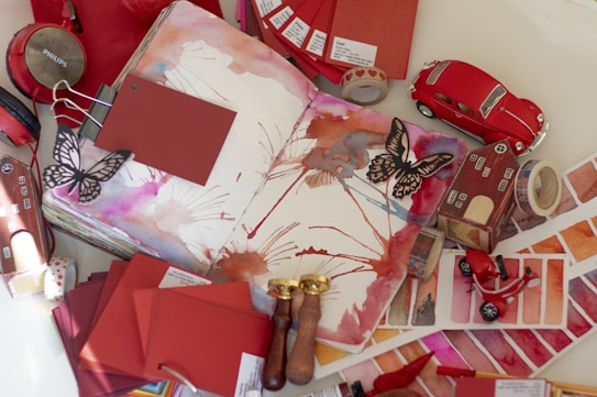 An open sketchbook features watercolor art with splashes of red, orange, and pink hues. Surrounding the sketchbook are various art and lifestyle objects, including red headphones, paint color swatches, a miniature red car, butterfly cutouts, paint swatches, a model house, and brass wax seals.