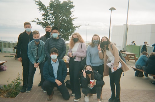A group of young people are gathered outdoors, wearing casual clothes and face masks. They appear to be posing for a picture with a relaxed and friendly demeanor. Some are standing while others are crouching, and there is a tree and park benches in the background. The setting seems to be a public park or outdoor area.