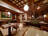 A cozy restaurant interior with wooden furnishings and soft lighting. The ceiling features a grid pattern with hanging spherical lights. Tables and chairs are neatly arranged, and a green cushioned bench lines the walls. A colorful floral painting adds vibrancy to the space.