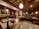 A cozy restaurant interior with wooden furnishings and soft lighting. The ceiling features a grid pattern with hanging spherical lights. Tables and chairs are neatly arranged, and a green cushioned bench lines the walls. A colorful floral painting adds vibrancy to the space.