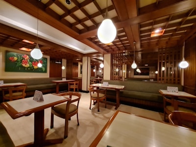 A cozy restaurant interior with wooden furnishings and soft lighting. The ceiling features a grid pattern with hanging spherical lights. Tables and chairs are neatly arranged, and a green cushioned bench lines the walls. A colorful floral painting adds vibrancy to the space.