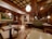 A cozy restaurant interior with wooden furnishings and soft lighting. The ceiling features a grid pattern with hanging spherical lights. Tables and chairs are neatly arranged, and a green cushioned bench lines the walls. A colorful floral painting adds vibrancy to the space.