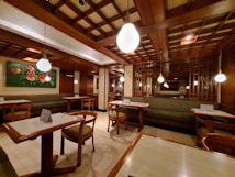 A cozy restaurant interior with wooden furnishings and soft lighting. The ceiling features a grid pattern with hanging spherical lights. Tables and chairs are neatly arranged, and a green cushioned bench lines the walls. A colorful floral painting adds vibrancy to the space.