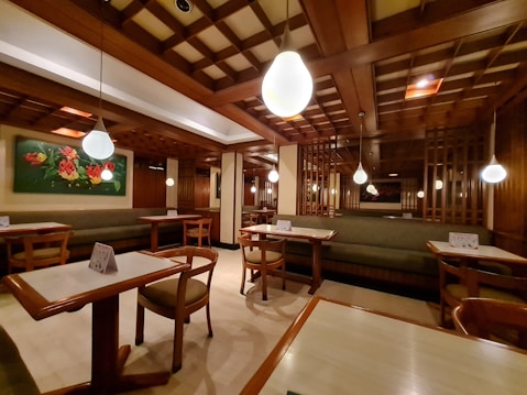 A cozy restaurant interior with wooden furnishings and soft lighting. The ceiling features a grid pattern with hanging spherical lights. Tables and chairs are neatly arranged, and a green cushioned bench lines the walls. A colorful floral painting adds vibrancy to the space.